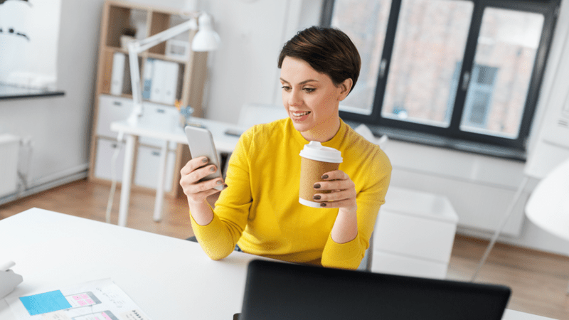 Woman in a yellow sweater looking at her phone while drinking a cup of coffee.