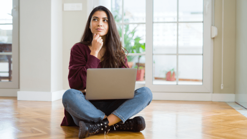 Woman sitting on the floor with her laptop holding her hand on her chin.