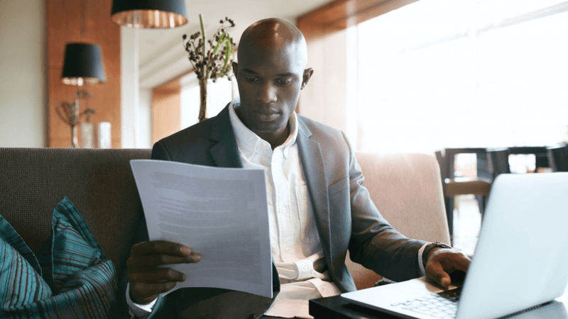 Man reading a document while typing on a laptop computer.
